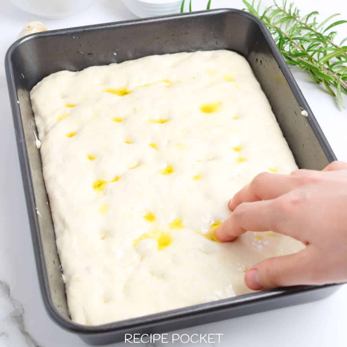 Close up of focicca dough in a black baking tin with a hand in the picture.