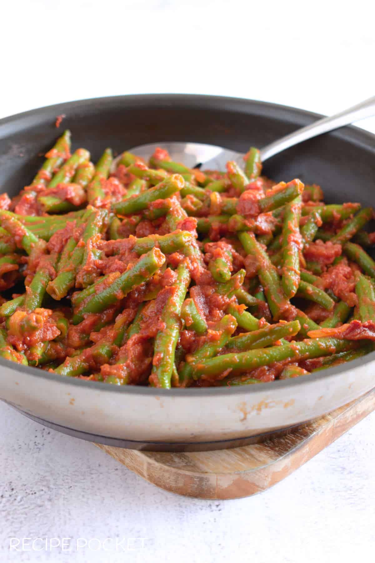 A frying pan filled with cooked green beans in tomato sauce with a serving spoon.