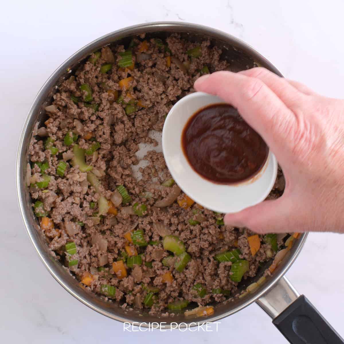 A hand holding tomato paste in a small bowl over a pot of cooked ground beef.