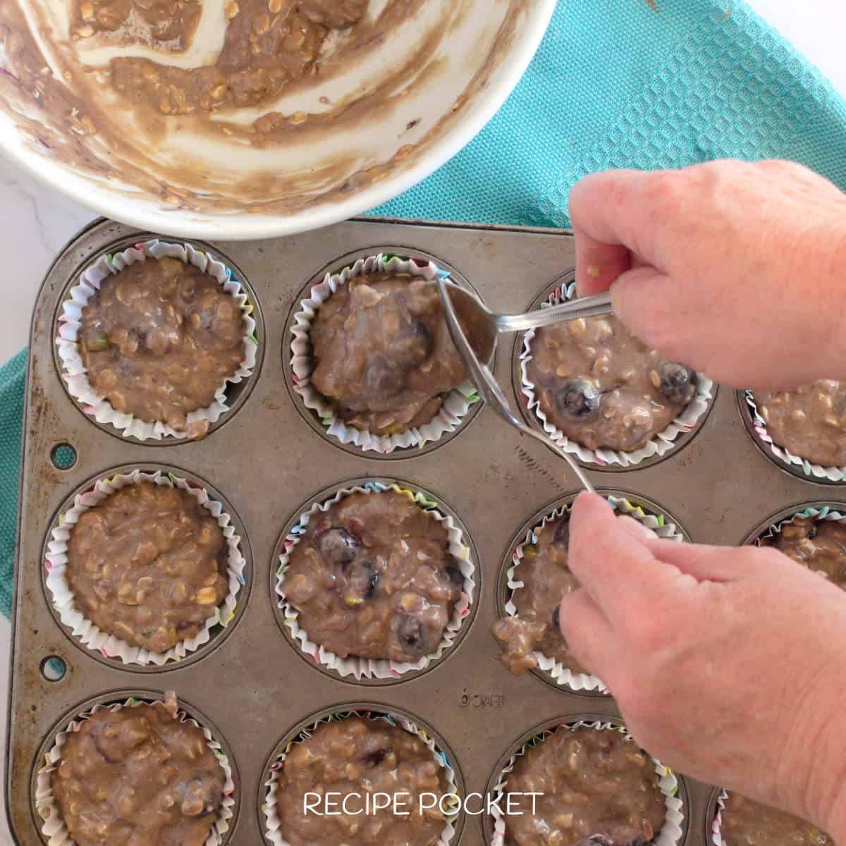 Muffin batter being spooned into paper cases in a muffin tin.